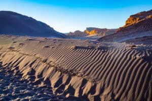 Desert landscape with rippled sand dunes and rugged mountains in the background.