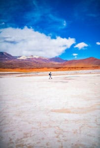 A person walking across a vast, arid desert with colorful mountains and a bright blue sky overhead.